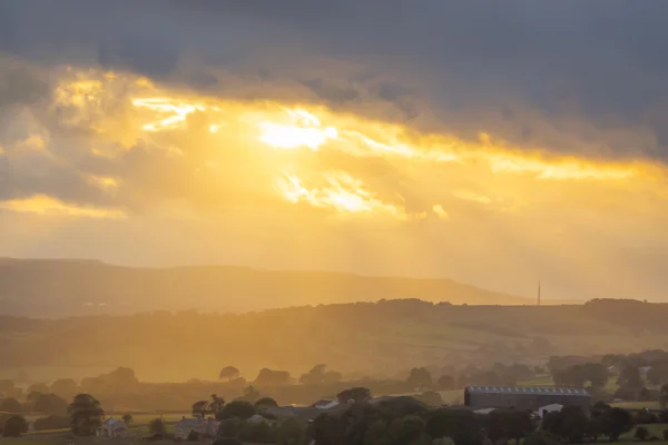 Sunbeams breaking through stormy cloud over a hazy moorland valley with farm buildings at dusk