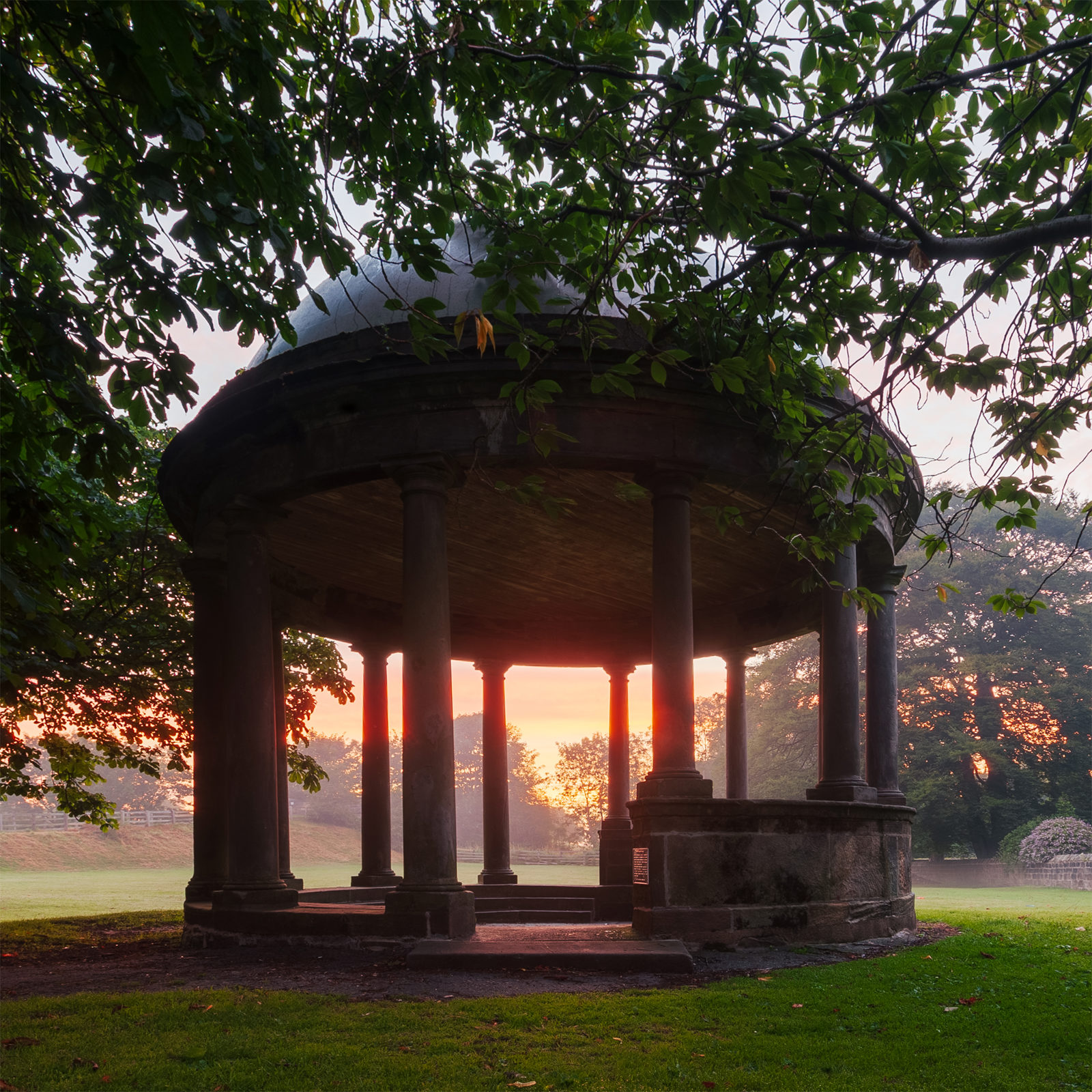 Circular stone pavilion with a domed roof supported by columns, surrounded by lush green grass and trees. The sky in the background displays a gradient of warm orange and pink hues, indicating a sunset or sunrise. Tree branches frame the pavilion, adding a natural, serene atmosphere.