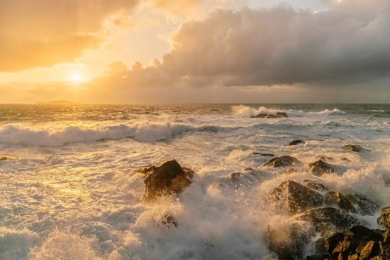 Sunset over a turbulent ocean with waves crashing against rocky formations. The sky is filled with dramatic clouds, illuminated in shades of orange and gold by the setting sun. A distant landmass is silhouetted on the horizon.
