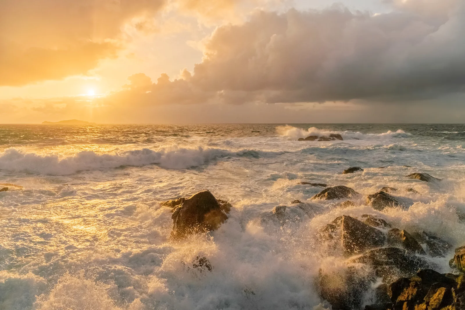 Waves crashing over rocks on a rugged coast at sunset, with golden light breaking through stormy clouds