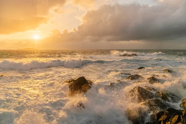 Waves crashing over rocks on a rugged coast at sunset, with golden light breaking through stormy clouds