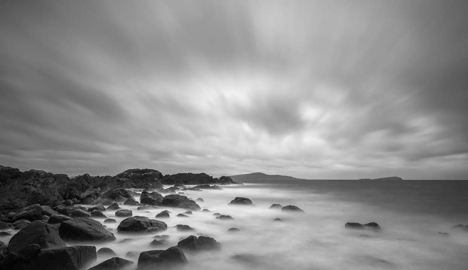Rocky shoreline with sea water blurring over the rocks, creating a misty effect. The sky is overcast with streaking clouds, suggesting a long exposure. A distant landmass is visible on the horizon. The image is in black and white, highlighting textures and contrasts.
