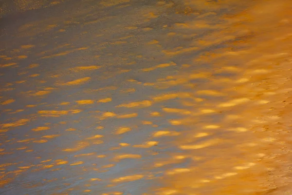 Shallow water with golden-brown mineral deposits and ripple patterns creating striped reflections.