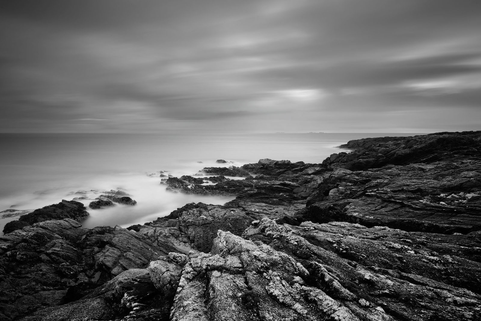 Dramatic black and white coastal landscape with rugged, weathered rocks in the foreground. The ocean appears misty and ethereal due to long exposure, blending with the soft, blurred sky. Gentle waves lap against the rocky shore, creating a serene atmosphere.