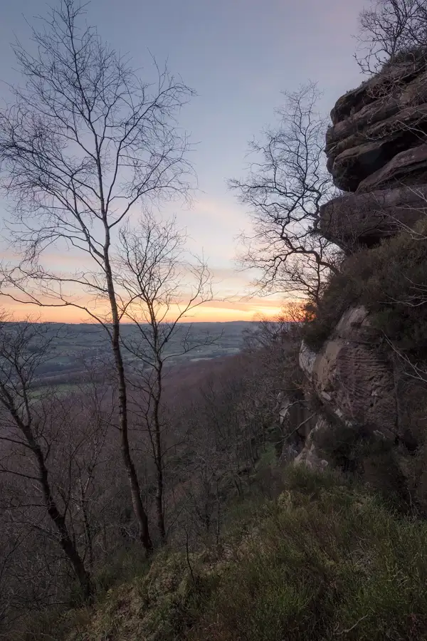 Bare trees stand on a steep hillside, overlooking a vast landscape of rolling hills and fields. The foreground features rocky outcrops on the right. The sky is painted in soft hues of orange and blue as the sun begins to set, casting a warm glow over the scene.
