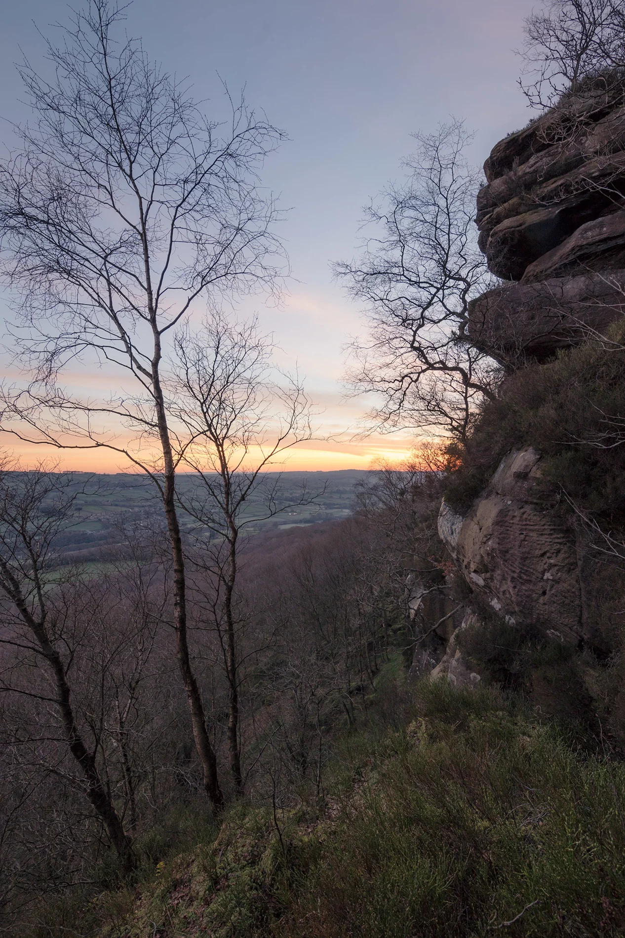 Sandstone outcrop and bare birch trees framing a hillside view towards a glowing winter sunset over patchwork farmland.