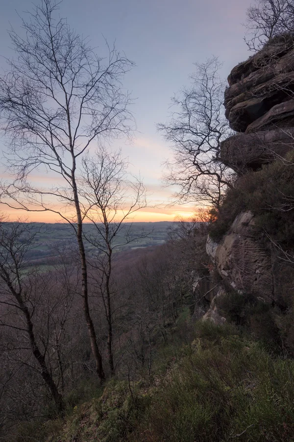 Sandstone outcrop and bare birch trees framing a hillside view towards a glowing winter sunset over patchwork farmland.
