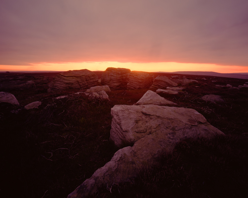 Moorland Landscapes: North Yorkshire moor at dusk. Large rocks and tufts of grass are silhouetted against the warm glow of a setting sun on the horizon, casting vivid hues of purple and orange across the sky. The scene conveys a tranquil, serene end to the day. a rocky landscape with a sunset