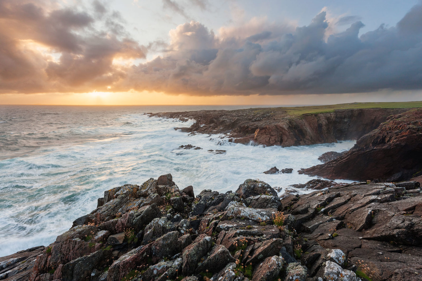 Rocky coastline with waves crashing against cliffs under a dramatic, cloudy sky. The sunset casts a warm golden light on the scene, creating a contrast with the dark rocks and turbulent sea. Some vegetation is visible on the rocks in the foreground.