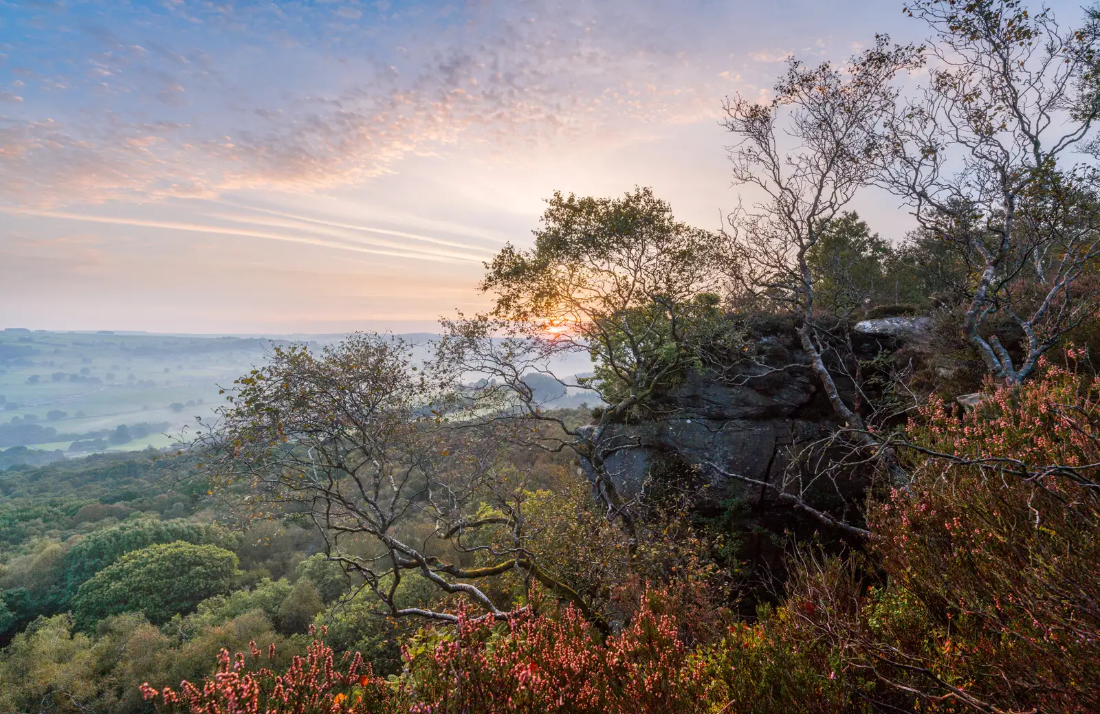 Sunset over a lush valley with rolling hills in the background. The foreground features a rocky outcrop and trees with sparse leaves, silhouetted against the sky. The sky is filled with streaks of orange and pink clouds. Pink heather is visible in the lower foreground.