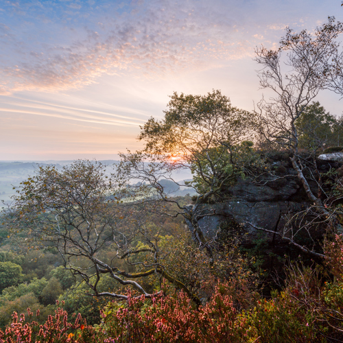 Edge of autum: Sunrise illuminates a countryside vista from a high vantage point. Various hues of pinks and blues adorn the sky. In the foreground are rocky outcrops and silhouettes of twisted trees. Below, layers of greenery blanket rolling hills, hinting at a tranquil rural landscape.