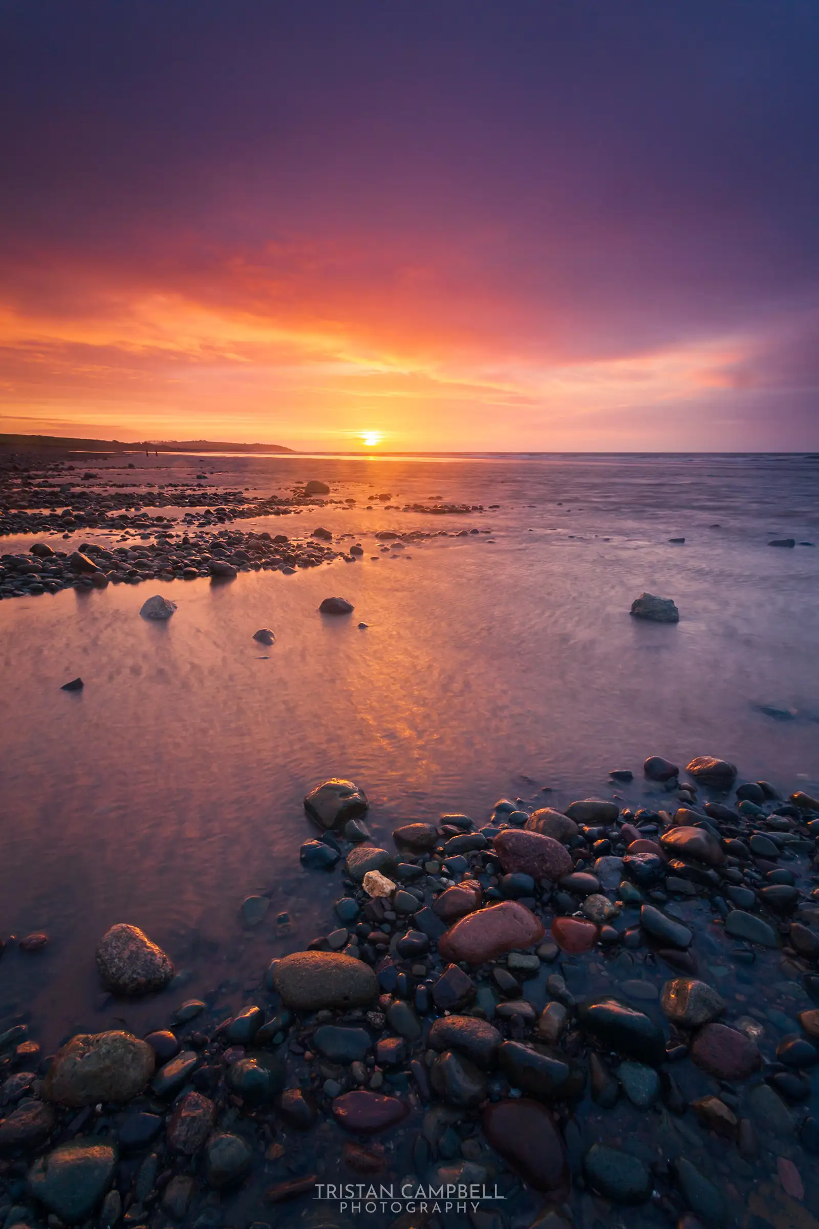 Sunset over a rocky beach with smooth stones scattered in the foreground. The sky displays a vibrant gradient of orange, pink, and purple hues, reflecting on the calm sea. In the distance, land forms a silhouette against the setting sun on the horizon.