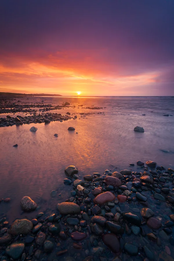 Pebble-strewn coastline at sunset with vivid orange and purple sky reflected in calm shallow water