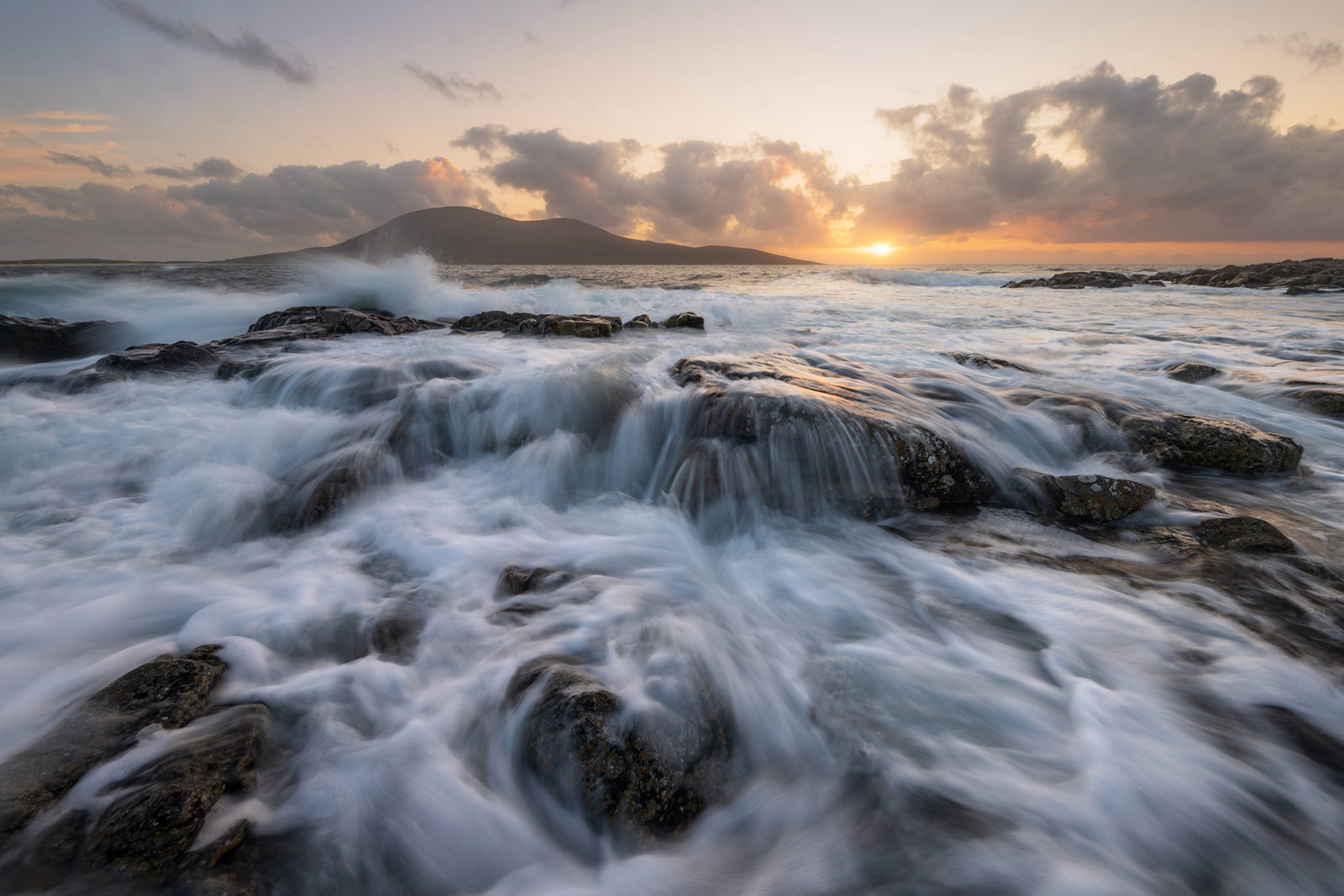 Waves crashing over dark rocks in the foreground create a sense of motion, with water appearing misty due to the long exposure. In the background, a silhouetted hill stands beneath a sky filled with dramatic clouds, as the sun sets on the horizon, casting a warm glow across the scene.