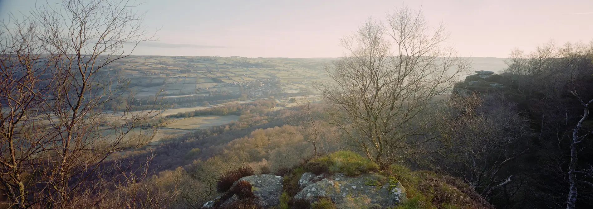 Patchwork valley of frosty fields and woodland seen from a gritstone edge with bare trees at dawn