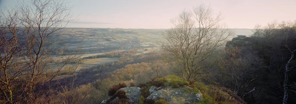 Patchwork valley of frosty fields and woodland seen from a gritstone edge with bare trees at dawn