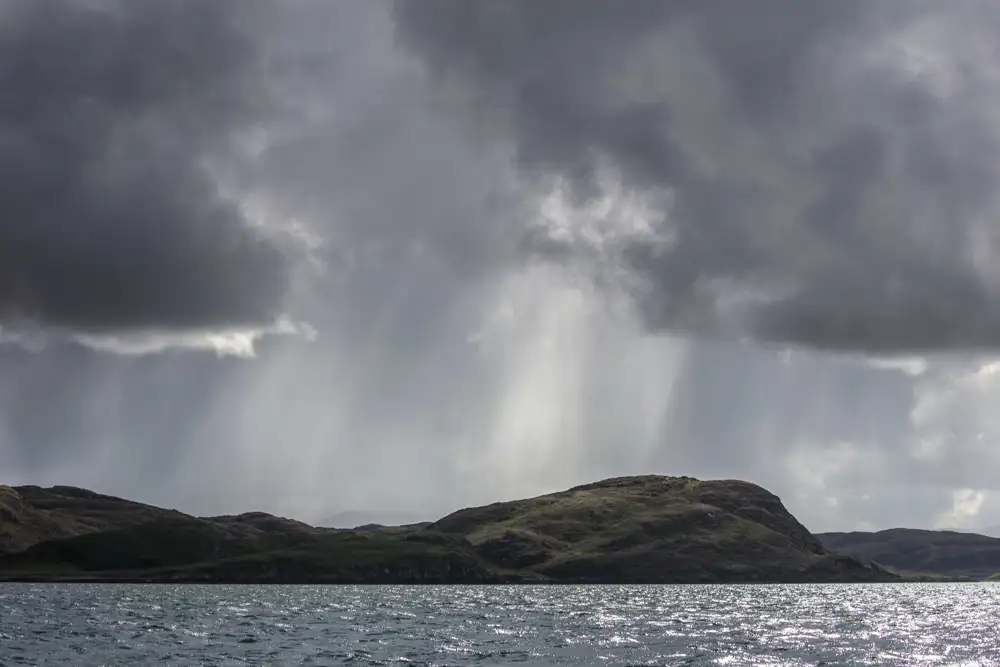 Dramatic grey clouds loom over a dark, rugged hillside by the edge of a shimmering body of water. Sunlight breaks through in rays, highlighting the contours of the landscape beneath the moody sky.