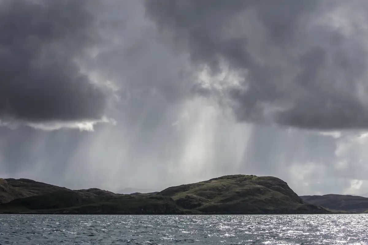 Dramatic grey clouds loom over a dark, rugged hillside by the edge of a shimmering body of water. Sunlight breaks through in rays, highlighting the contours of the landscape beneath the moody sky.