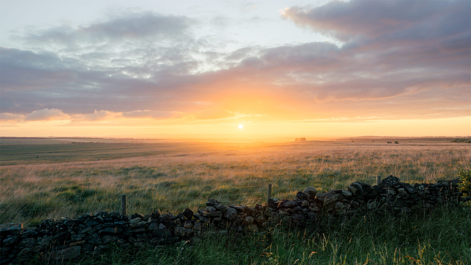 Sunset over a vast, grassy field with a low, dry stone wall in the foreground. The sky is filled with clouds, mostly grey but tinged with warm hues from the setting sun, which casts a golden light across the landscape.