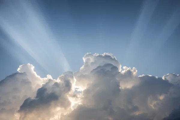 Sunbeams bursting through towering cumulus clouds against a clear blue sky