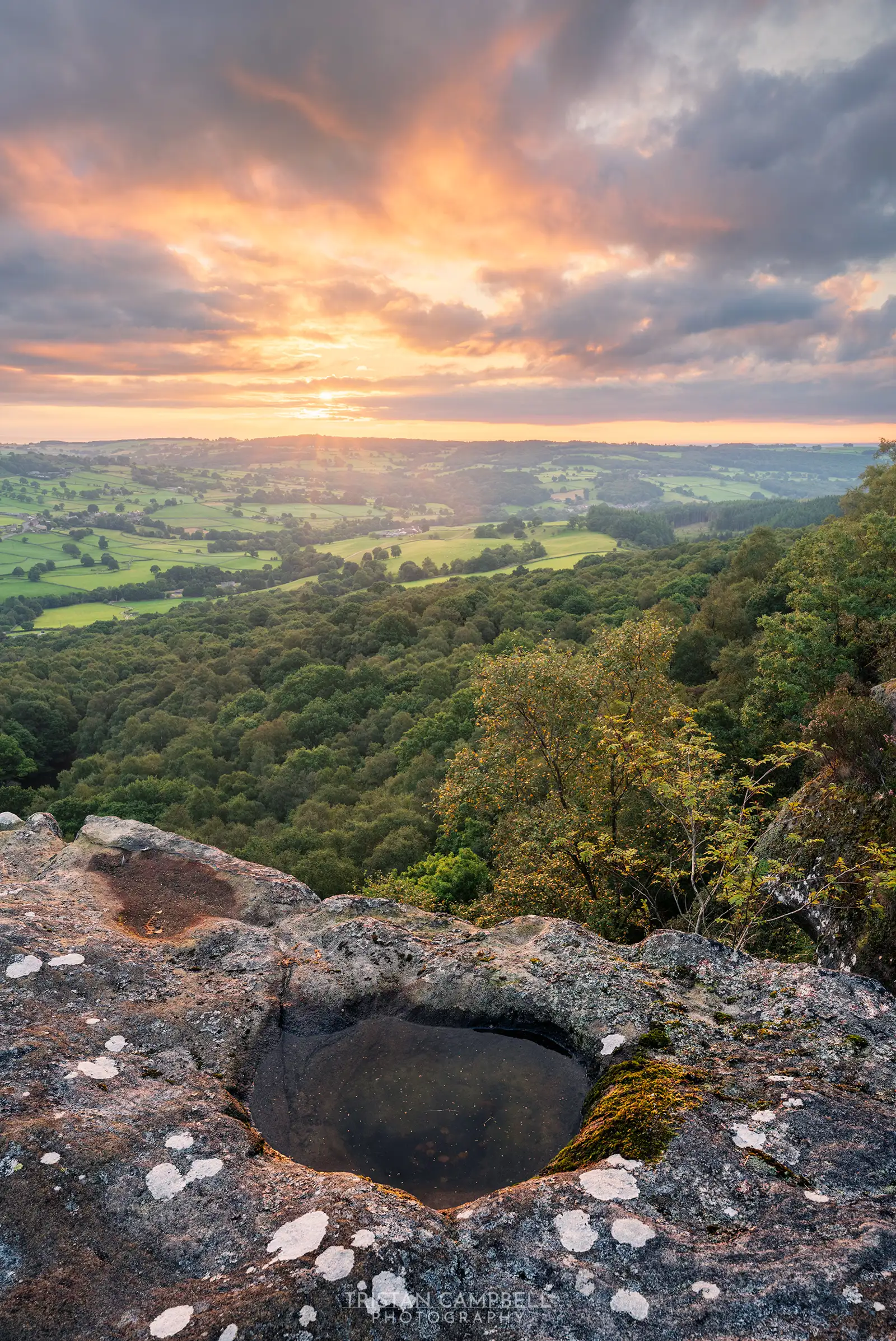 Dramatic sunset over a lush, rolling landscape viewed from a rocky cliff with a small pool of water reflecting the sky. The foreground shows a rocky surface adorned with patches of lichen and moss. Beyond, expansive fields and dense forests stretch towards the horizon, bathed in warm orange and pink hues from the setting sun, partially obscured by fluffy clouds.