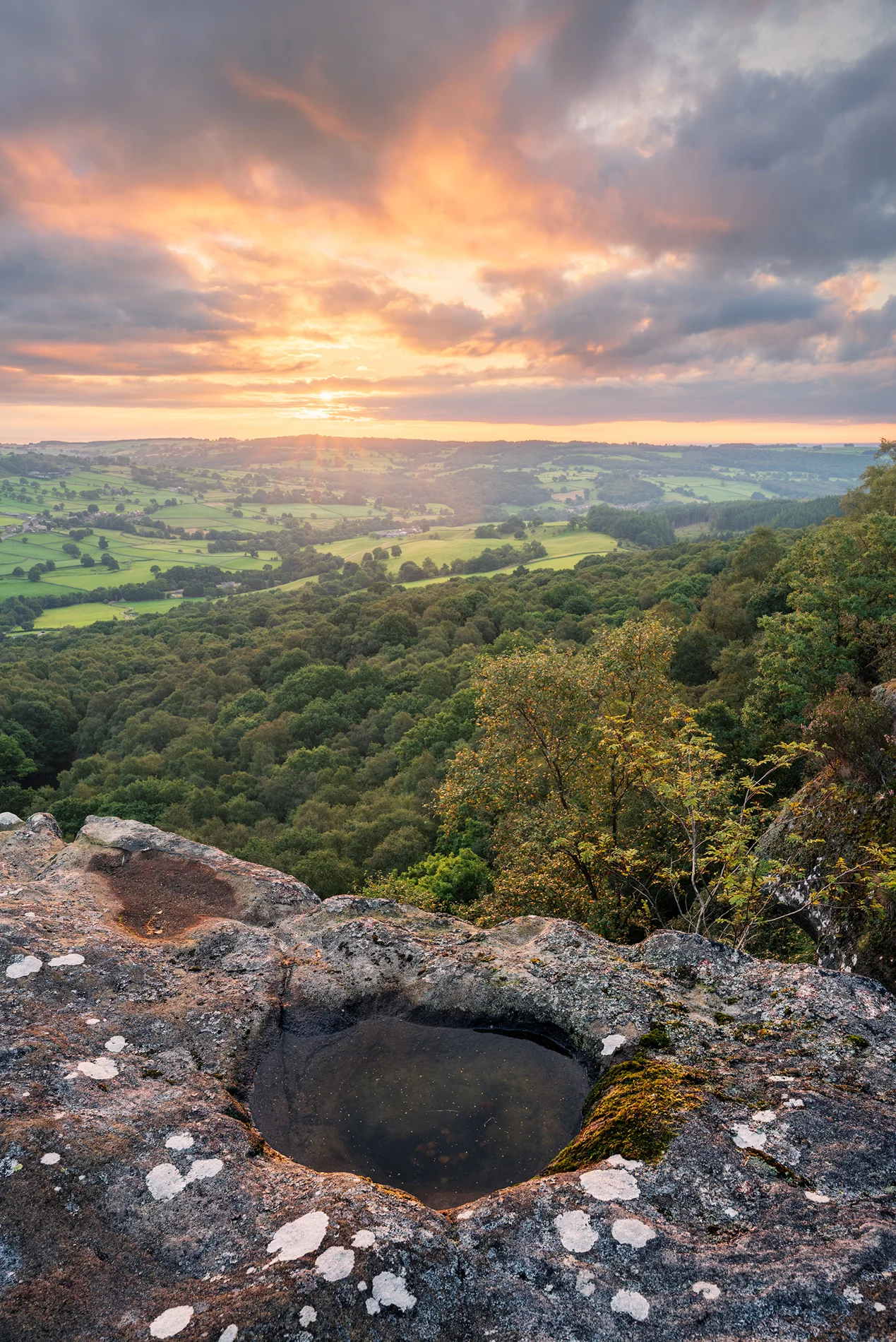Rock pool on a gritstone edge above a wooded valley and patchwork farmland at sunset