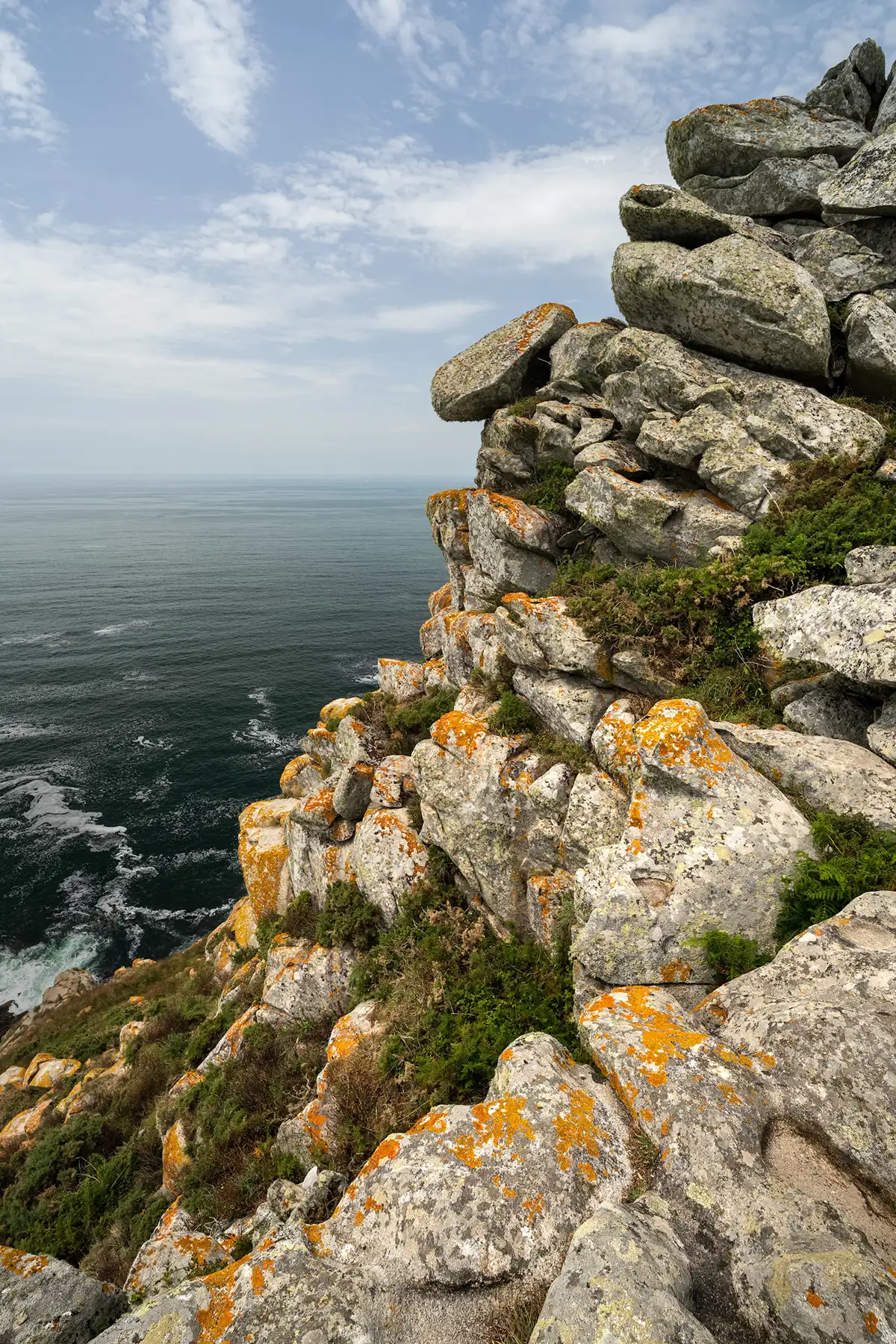 Rocky cliffside with large boulders covered in orange and yellow lichen, overlooking a vast, calm ocean under a partly cloudy sky. Vegetation grows among the rocks.