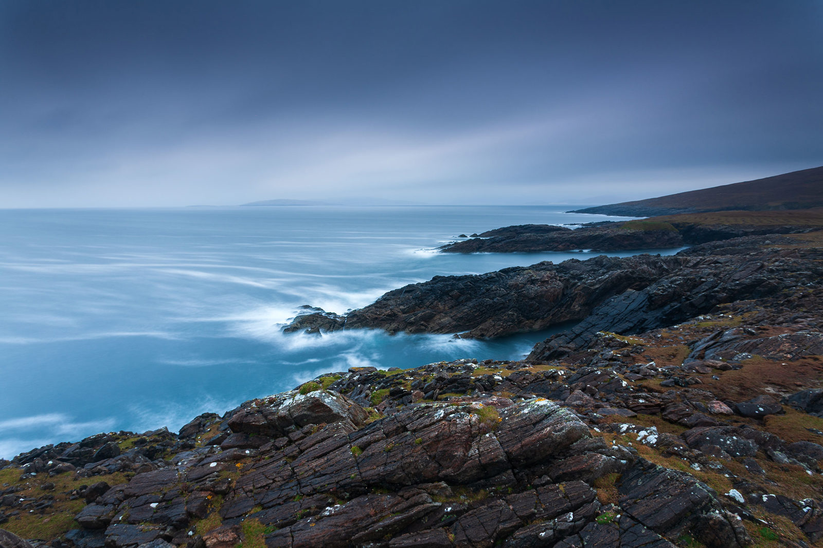Rocky coastline with rugged cliffs extending into a blue sea under a cloudy sky. The long exposure creates a smooth, misty effect on the water, contrasting with the dark, textured rocks. The horizon is faintly visible in the distance, while the landscape slopes gently upward to the right.
