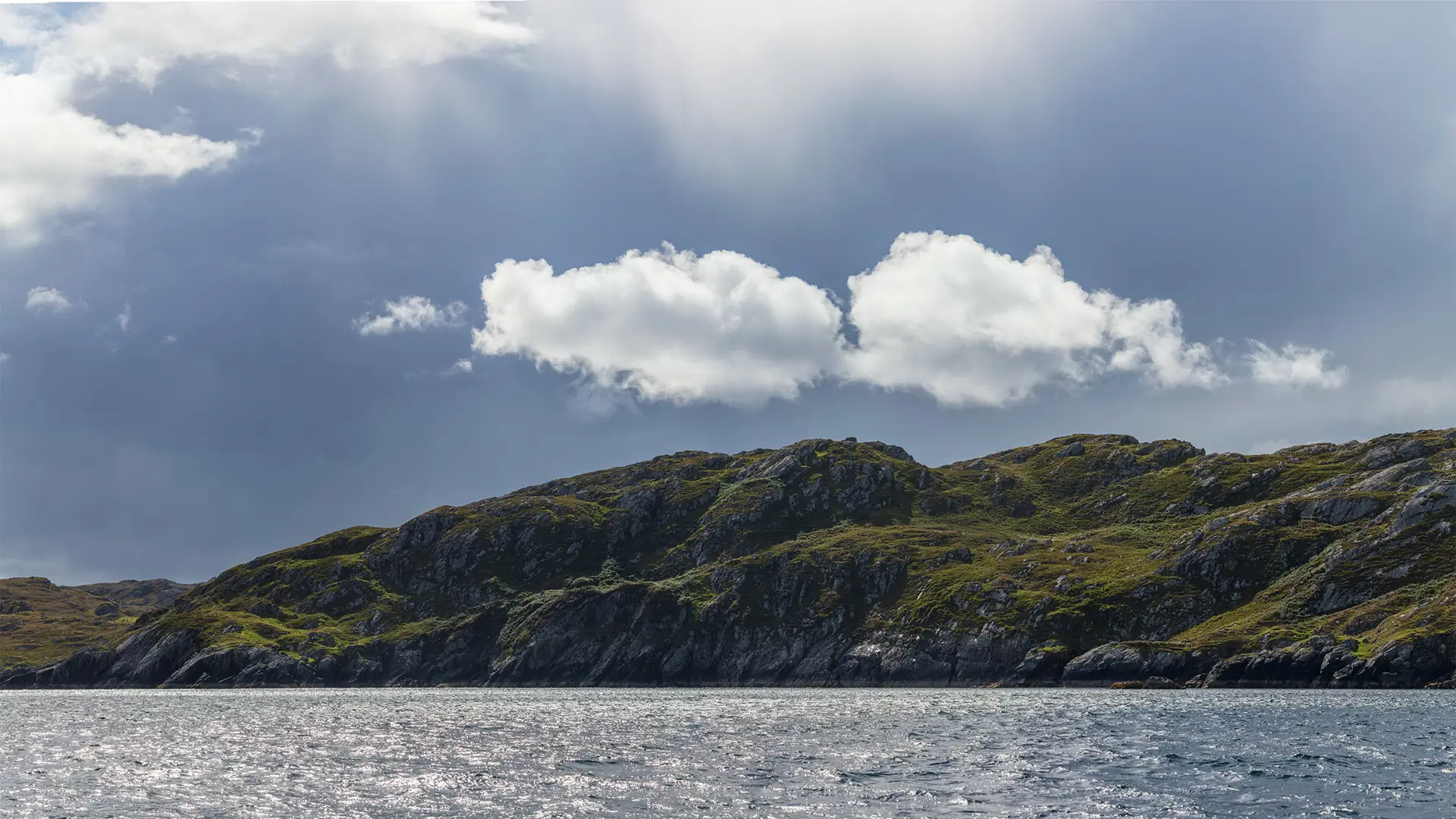 Rugged coastal hills covered with patches of grass and rock under a cloudy sky. The calm sea reflects the light, with waves gently lapping at the shoreline. Fluffy white clouds hover above, contrasting with darker clouds in the background.
