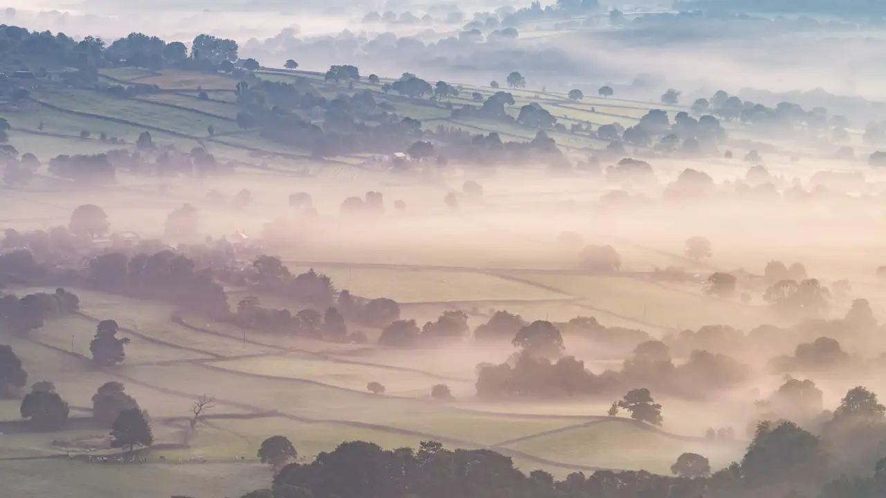 Rolling countryside landscape with patchwork fields partially obscured by a light mist. Trees dot the fields, creating a layered effect as the mist softens the contours. Soft morning light adds a serene and ethereal quality to the scene.