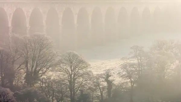 A misty scene with a stone viaduct in the background featuring multiple arches. Frosted trees with bare branches occupy the foreground, and a few sheep graze on the frosty grass below, creating a serene, wintry atmosphere. The light is soft and diffused through the mist, adding to the ethereal quality of the landscape.