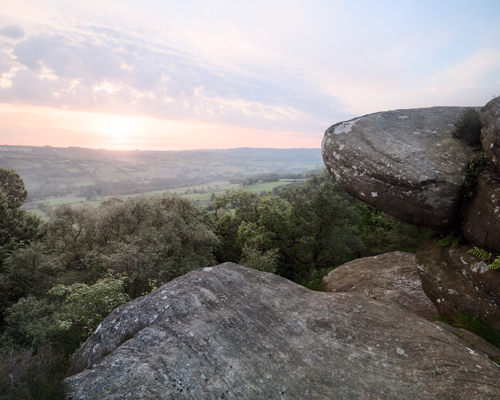 Moorland Landscapes: Large, weathered rock formations, covered with lichens, dominate the foreground while a lush canopy of trees spreads out below. The sky is painted with soft pastel hues as the sun dips near the horizon, casting a warm glow over the tranquil, rolling countryside. a view of a valley with trees and a sunset