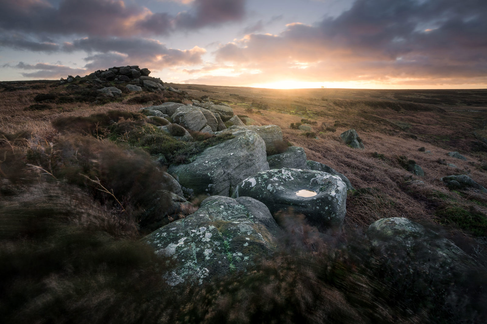 Rocky moorland landscape at sunset, with a skyline of dramatic clouds illuminated by warm light. Foreground features large boulders with patches of lichen and a small pool of water. Sparse grass and heather surround the rocks, creating a rugged and tranquil scene.