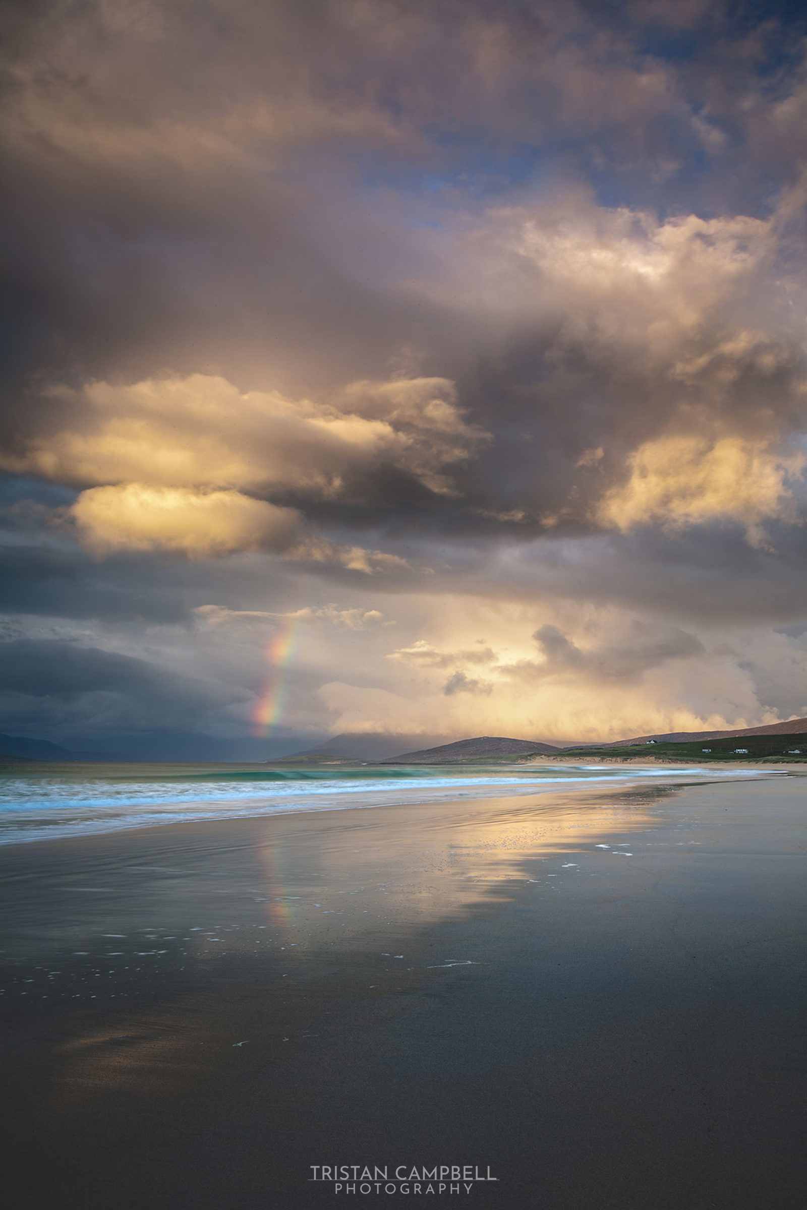 Dramatic, cloudy sky with golden light illuminating the clouds. A faint rainbow appears in the centre, partially reflected on the wet sand of a wide beach. Gentle waves wash onto the shore, and distant hills are visible under the moody sky.