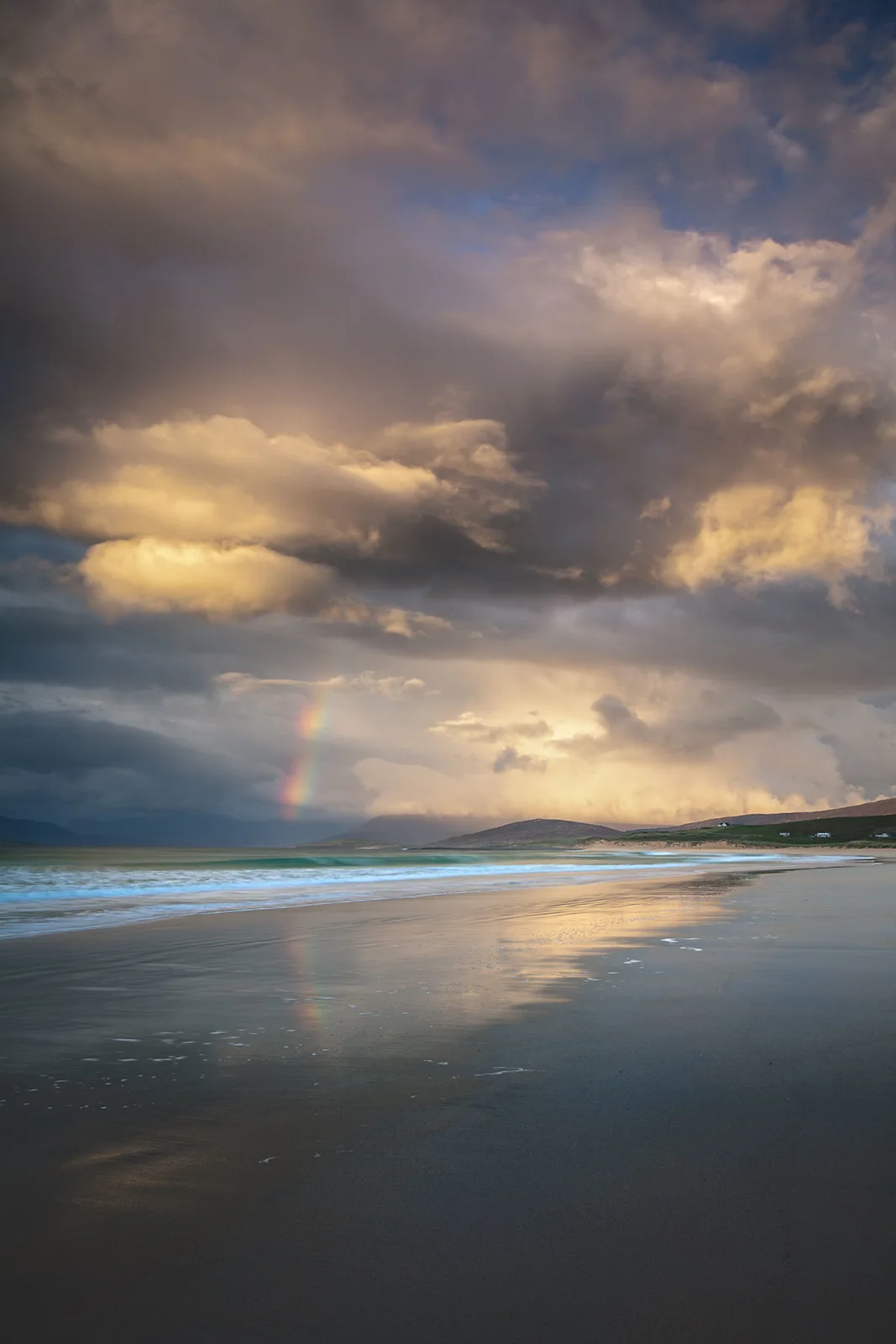 Wet sandy beach with turquoise waves, double rainbow, and dramatic cloudy sky with golden light over distant hills.