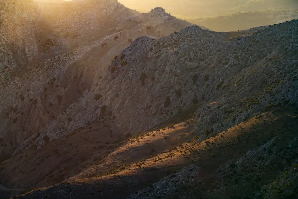 Rocky mountain landscape with soft, golden sunlight illuminating the slopes. Jagged cliffs and scattered shrubs are visible, and the sun casts long shadows across the terrain, highlighting textures and contours. The light fades into a more diffuse and misty background, accentuating the ruggedness of the scene.