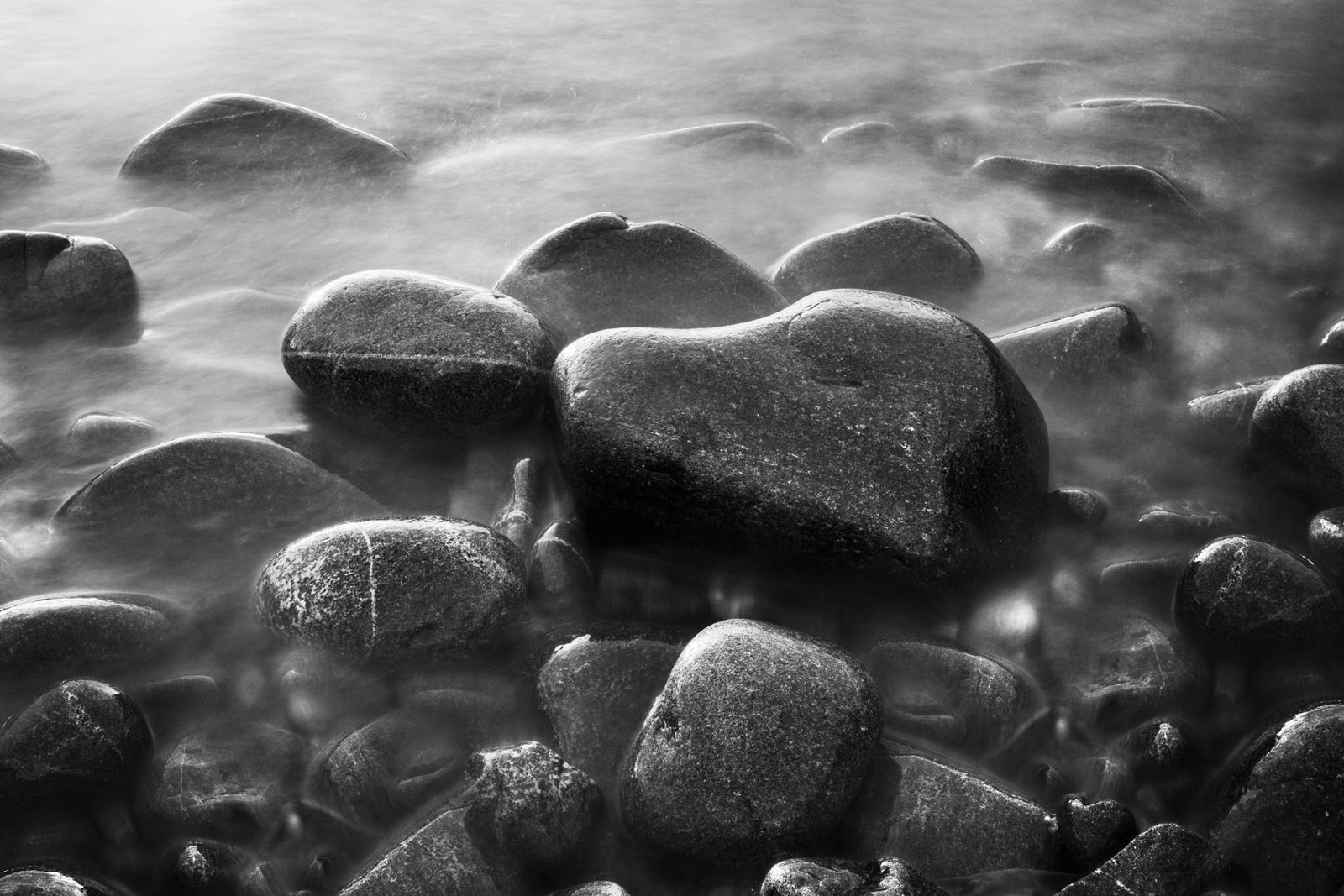 Smooth pebbles are clustered together, partly submerged in misty waters that create a serene atmosphere. The scene is captured in monochrome, accentuating contrasts and textures on the stone surfaces.