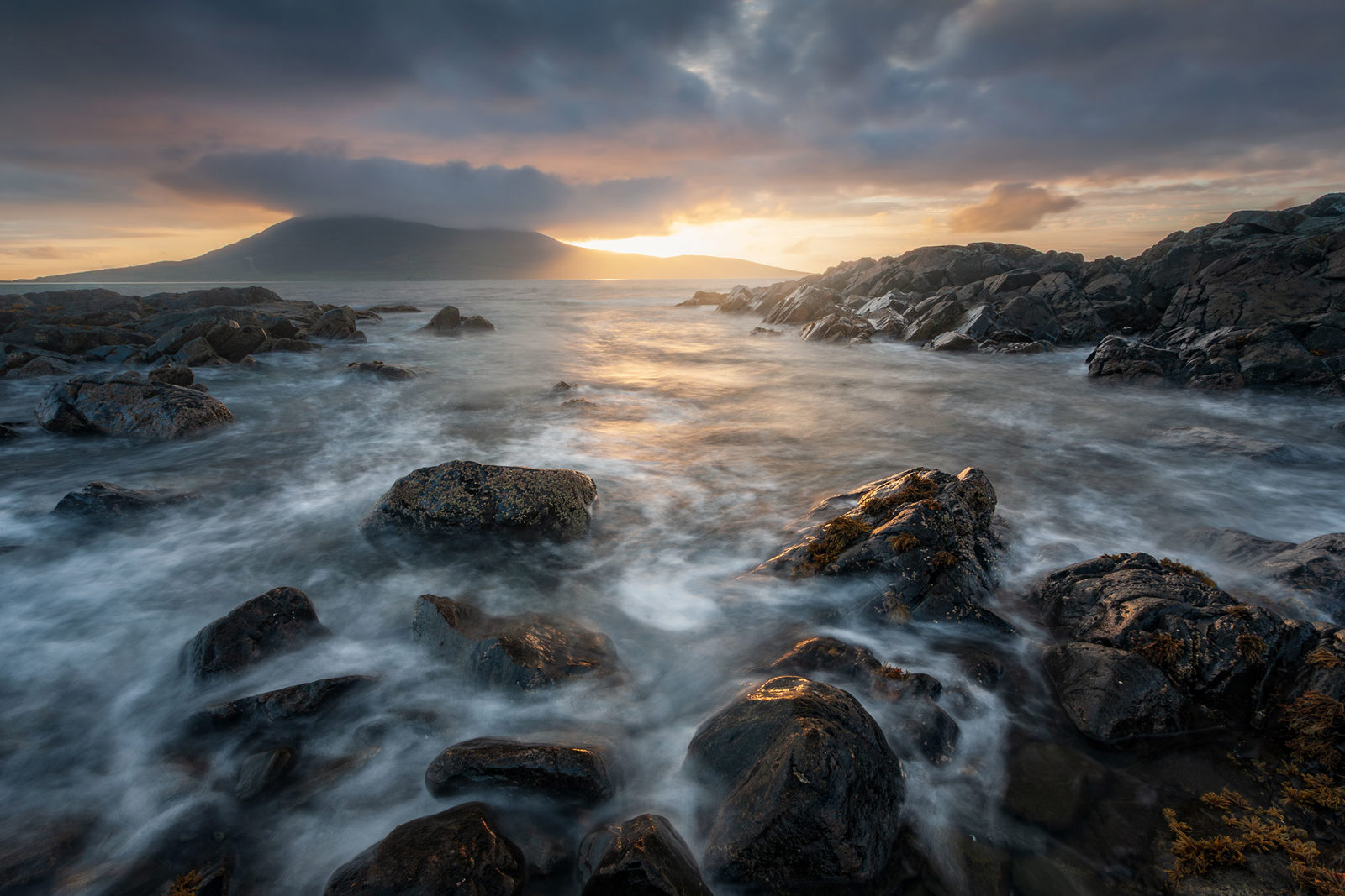Rocky shoreline with waves gently washing over the stones, under a cloudy sky at sunset. The horizon features a silhouette of a mountain partially covered by clouds, with warm sunlight breaking through and reflecting on the water's surface.