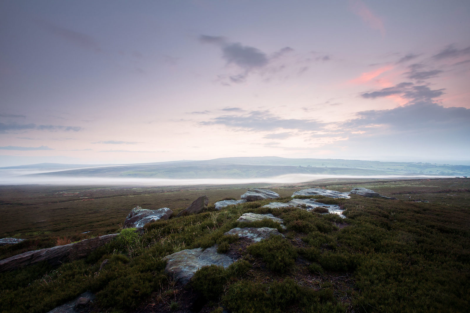 Moorland landscape at dawn with low-lying mist stretching across the scene. The foreground features rocks and patches of heather, leading to expansive moors. The horizon is softly lit in shades of pink and purple, with a few clouds scattered across the sky.
