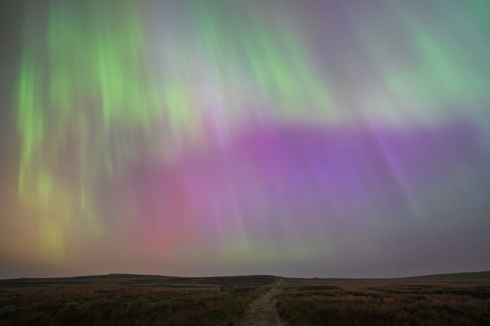 Aurora borealis dancing above rolling moorland with a dirt track, displaying vivid green, pink, and purple colours against a star-speckled night sky.