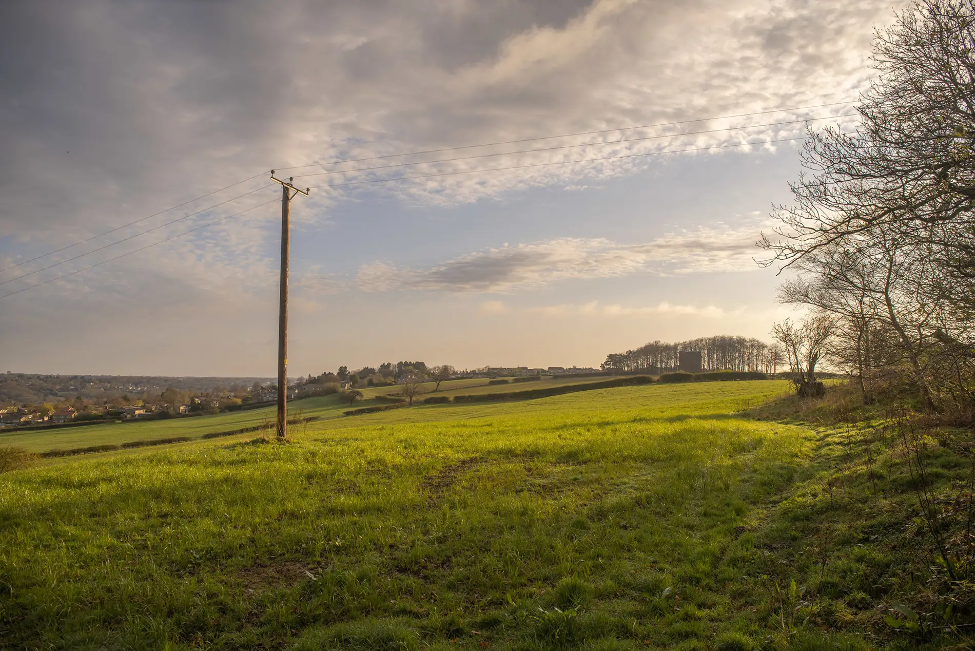 Telegraph pole in a green hillside field with a village in the valley below under a soft cloudy sky