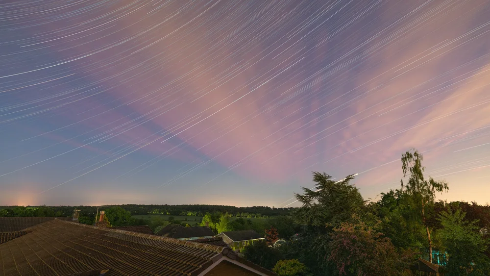 Star trails arc over a rural village rooftop and trees beneath a pink and blue twilight sky.