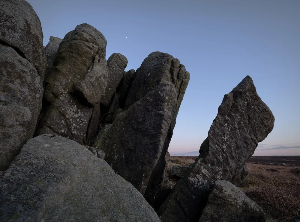 Lichen-covered gritstone tor on open moorland under a clear twilight sky with a faint moon