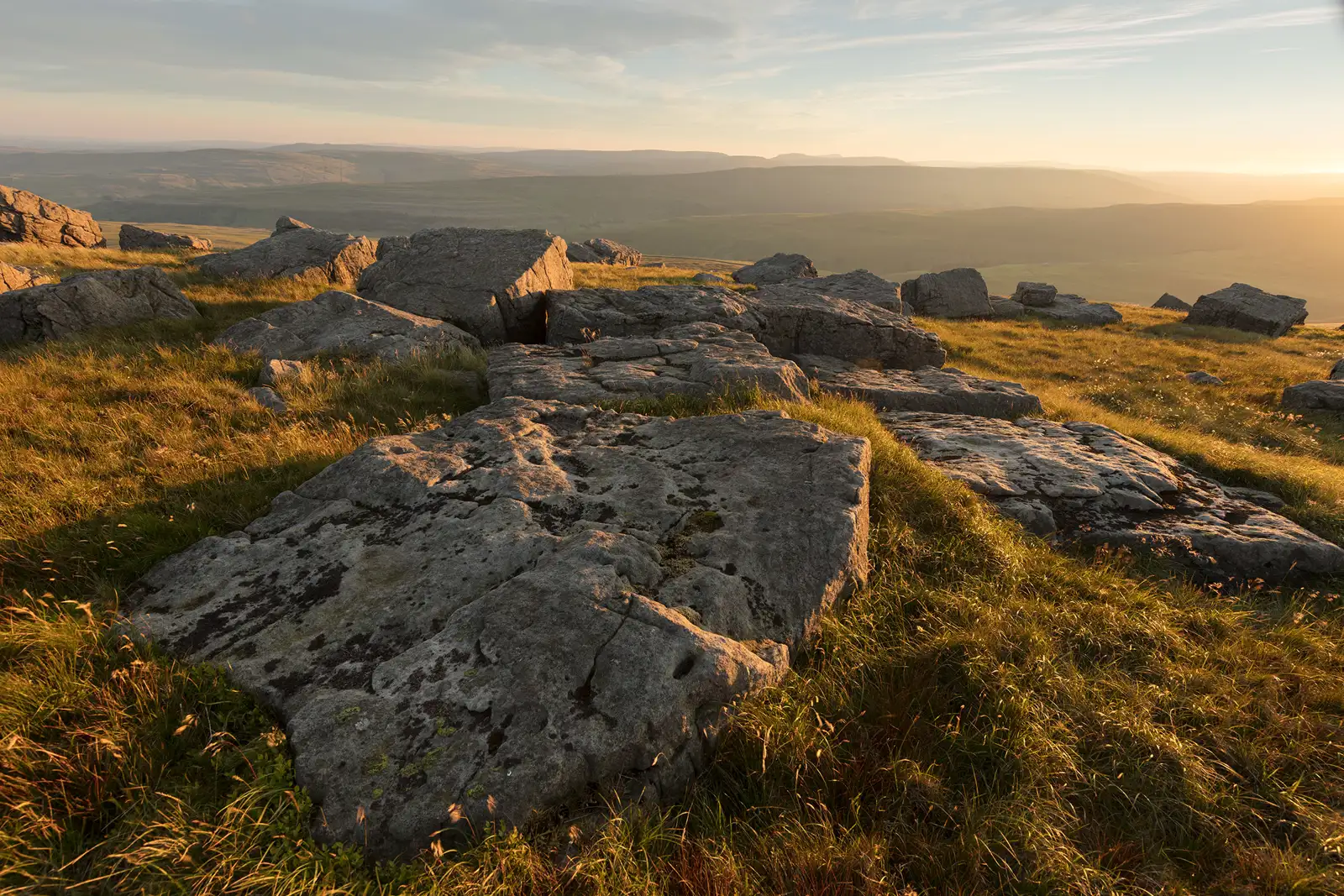 Rocky landscape bathed in sunset light, with large flat stones and patches of grass. The golden glow of the sun casts long shadows, and rolling hills are visible in the distance under a partly cloudy sky.