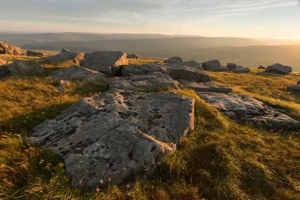 Rocky landscape bathed in sunset light, with large flat stones and patches of grass. The golden glow of the sun casts long shadows, and rolling hills are visible in the distance under a partly cloudy sky.