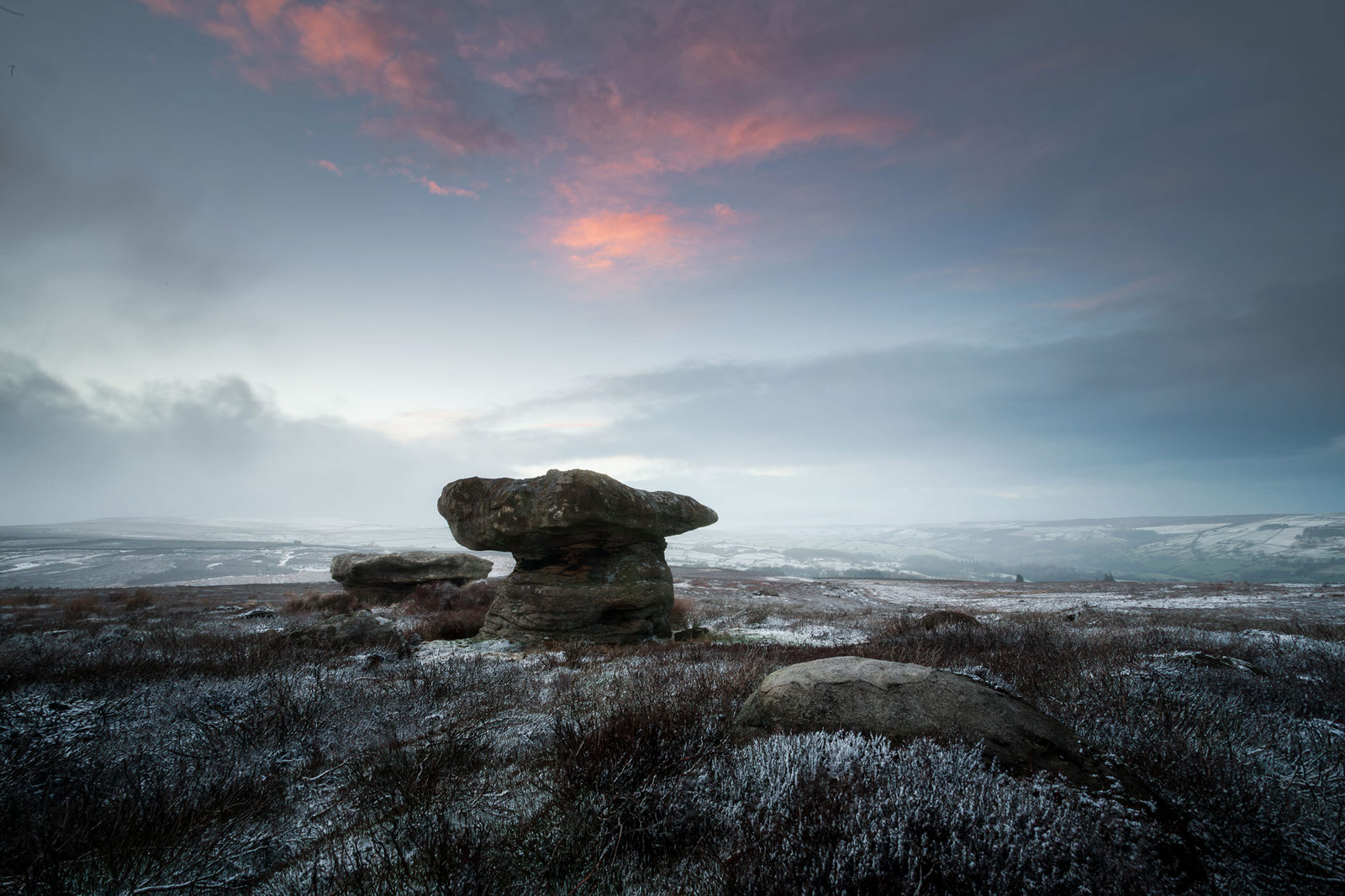 Snow-dusted moorland with large rock formations in the foreground, under a dramatic sky with soft pink clouds. The landscape extends into the distance with gentle hills and a pale horizon, creating a serene and cold winter atmosphere.