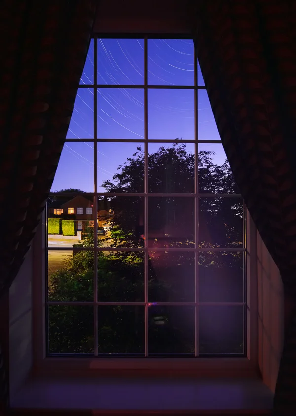 Star trails over a suburban street framed by a panelled sash window at twilight, with curtains drawn back