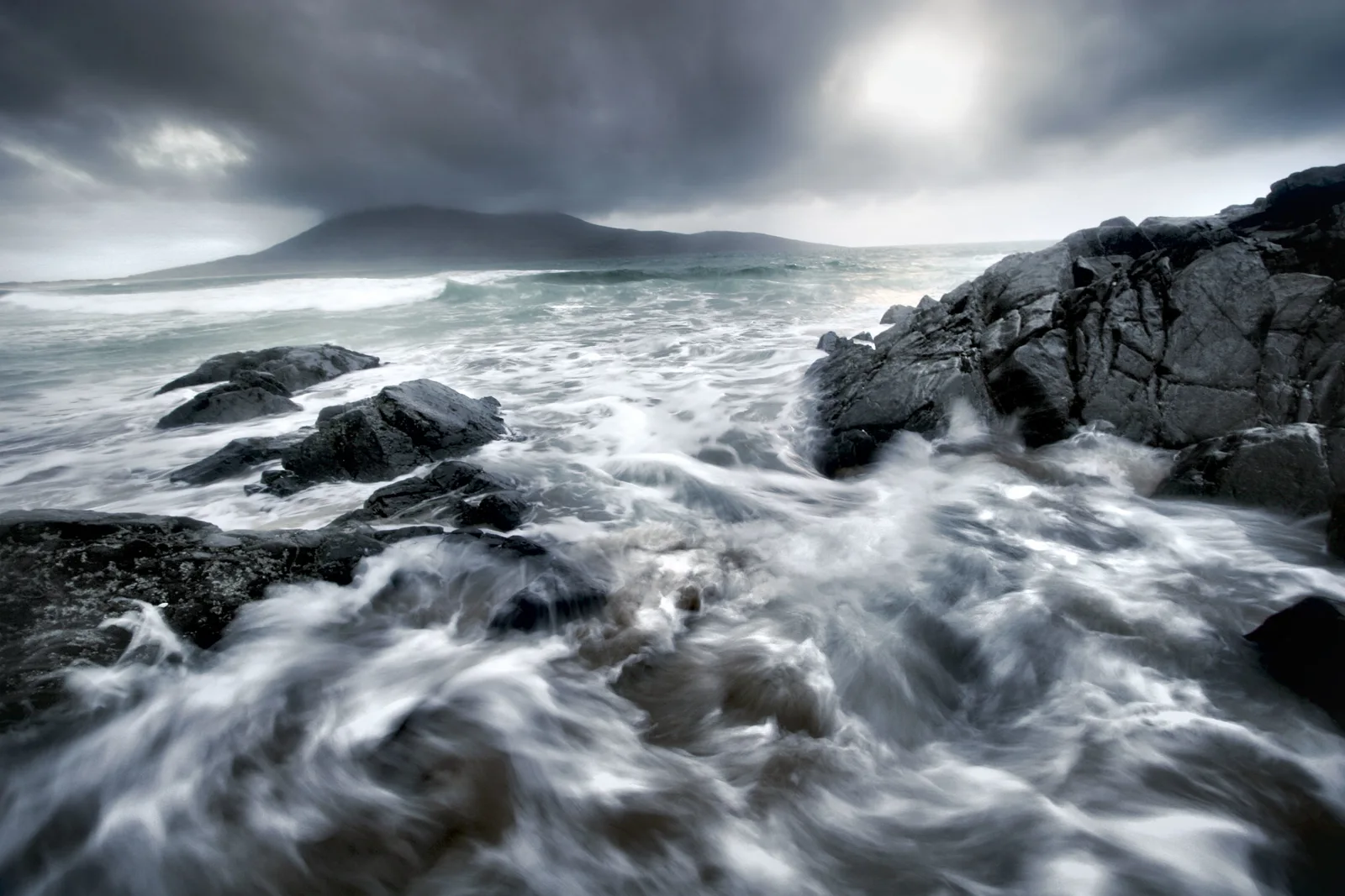 Stormy seascape with dark rocky outcrops, turbulent white waves, and a misty mountain in the distance under grey cloud cover.