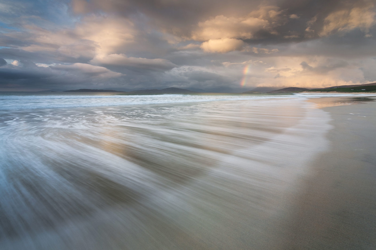 A beach scene with waves washing over the sand, creating a smooth, blurred effect. The sky is filled with dramatic clouds, illuminated by warm sunlight. A faint rainbow is visible in the distance near the horizon, with dark hills in the background.