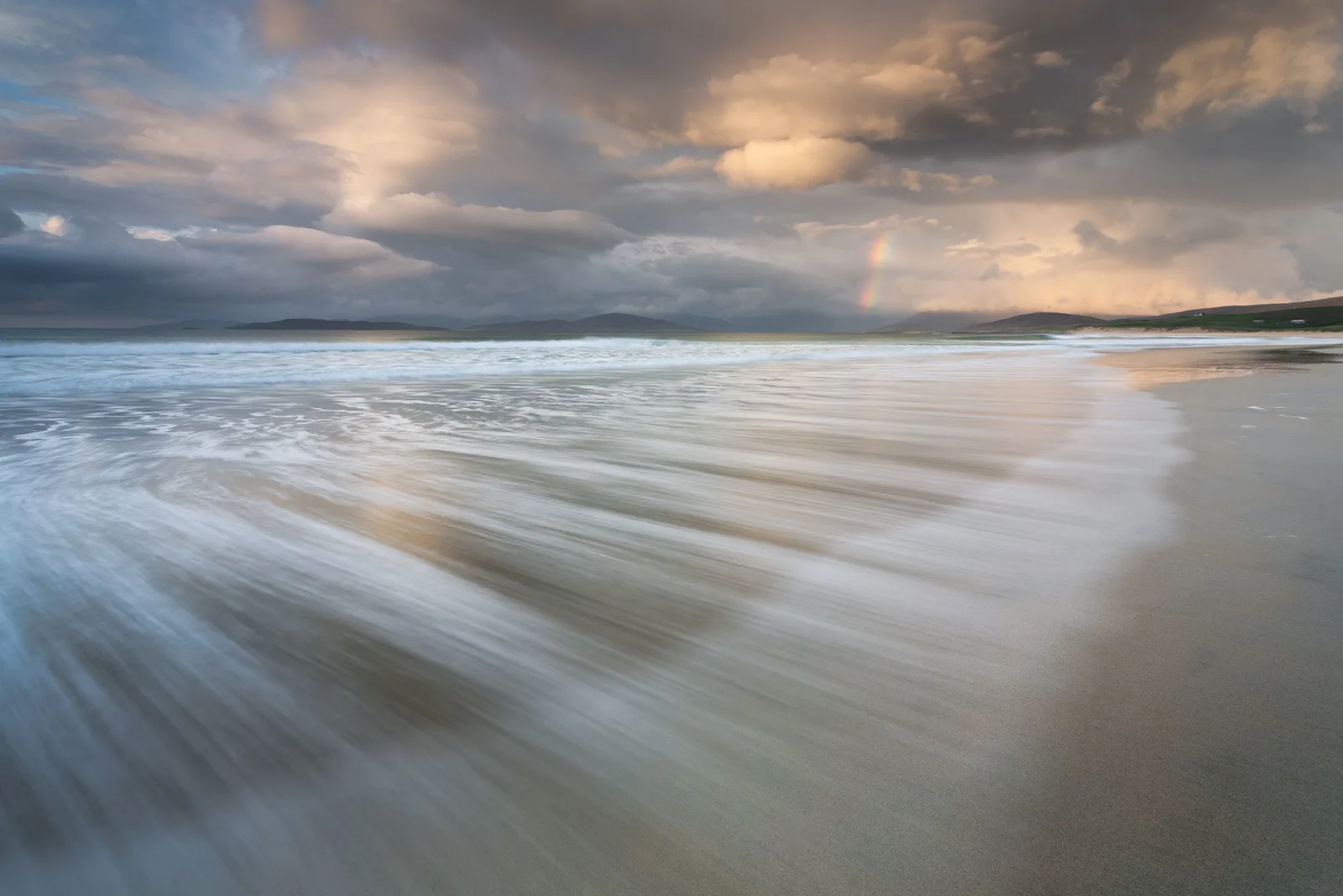 Sandy beach with rolling waves beneath dramatic cloudy sky and distant mountains; faint rainbow visible on horizon.